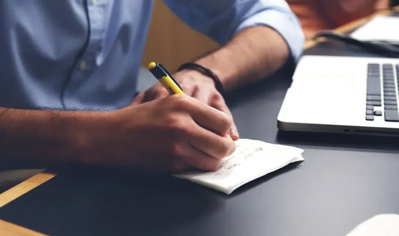 A person in a blue shirt is writing with a pen on a notepad next to a laptop on a black table.