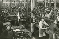Black-and-white photo of a busy factory floor with many workers operating machinery and assembling parts at workbenches.
