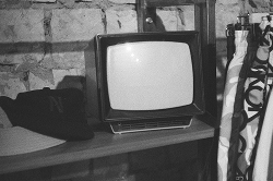 A small, vintage television sits on a wooden shelf against a stone wall, next to folded fabric and a dark object.