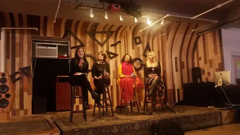 Four women sit on stools on a small stage with wood-paneled backdrops, participating in a discussion panel. Three hold microphones. Behind them, abstract black shapes adorn the wall. Stage lights and audio equipment are visible, and an AC unit is mounted on the left wall.