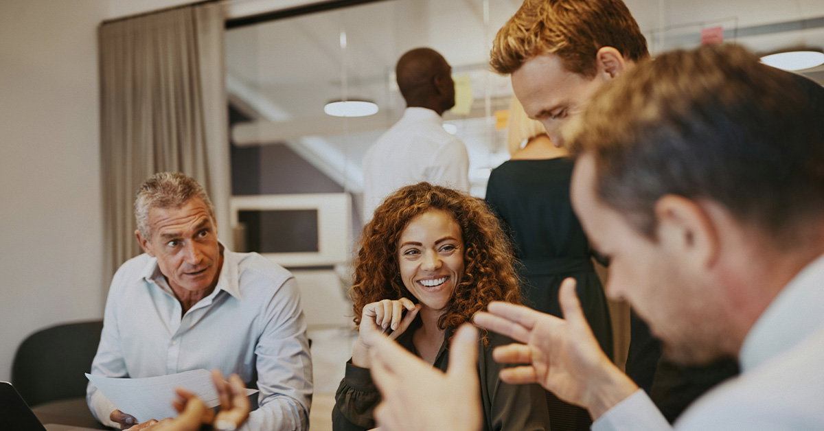 Four people are sitting around a table in an office, having a discussion about healthcare industry challenges. One woman is smiling while the others talk. Two more people are visible in the background.