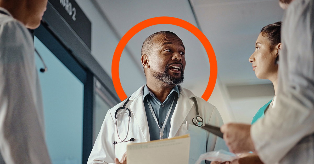 A doctor holding papers speaks with colleagues in a hospital hallway, an orange circle behind his head highlighting the teamwork essential to reducing employee turnover in healthcare.