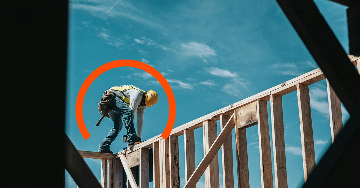 A construction worker in a yellow hard hat and safety vest bends down on a wooden framework under a blue sky, with an orange semicircle graphic behind them—capturing the hands-on reality of manufacturing industry challenges.