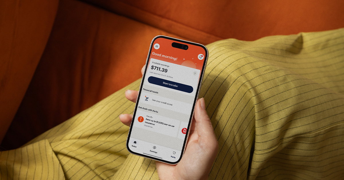 A hand holds a smartphone displaying an on-demand pay financial app with a balance of $711.39, set against a yellow pinstriped fabric and an orange surface.