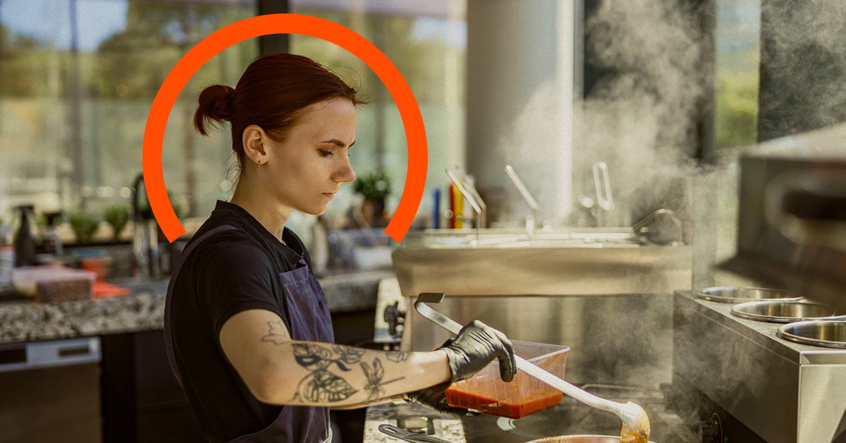 A person with tattoos and gloves ladles sauce in a commercial kitchen, with steam rising from pots and an orange arc graphic behind their head, reminiscent of the heat felt during inflation statistics 2022.
