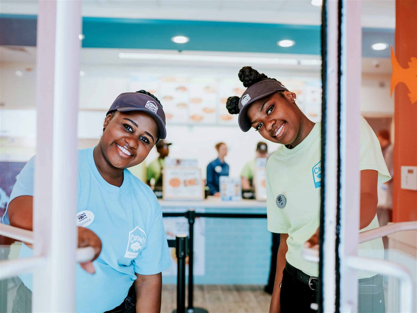 Two smiling employees in blue uniforms standing behind a counter in a brightly colored fast-food restaurant, one is opening the door for the viewer.