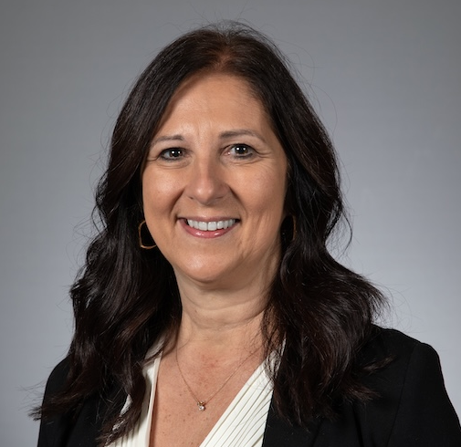 Woman with long dark hair wearing a black blazer and white blouse, smiling at the camera against a plain gray background.