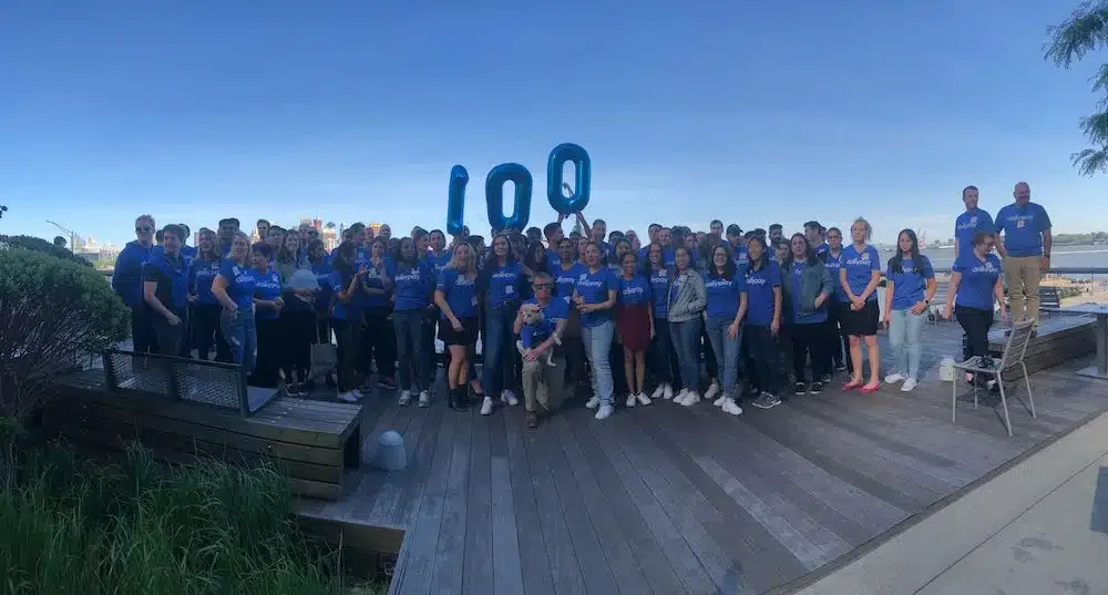 A large group of people wearing blue shirts gather on an outdoor wooden deck, posing with blue balloons forming the number "100"; trees and a distant water body are in the background.