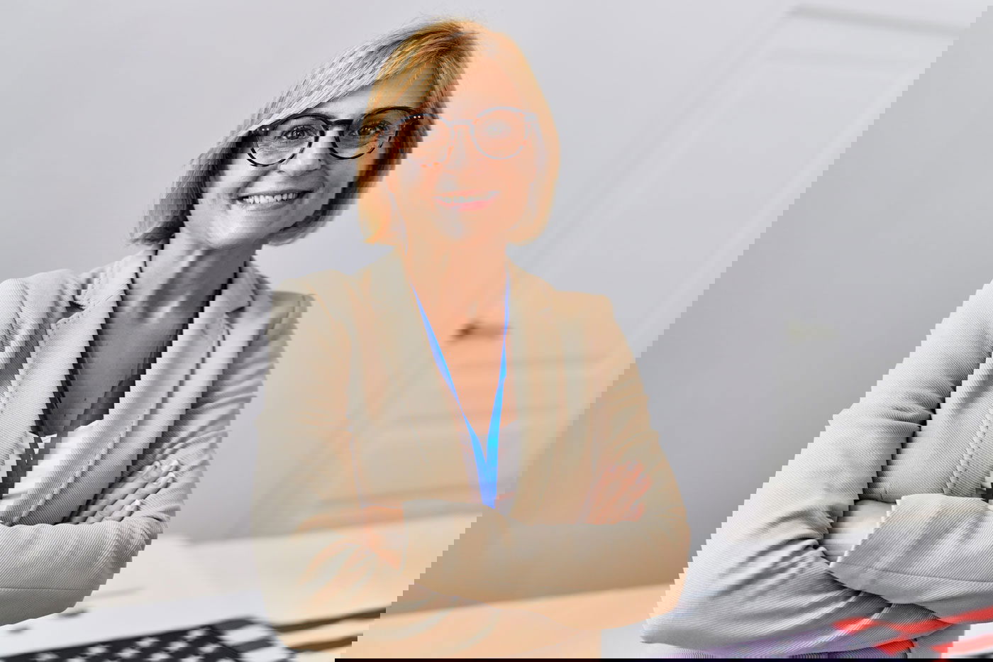 A woman with glasses and a beige blazer stands confidently with her arms crossed in front of a table with a partial view of an American flag.