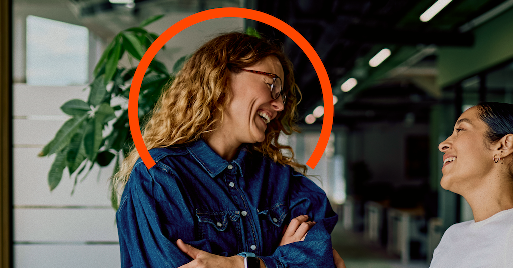 Two people stand indoors having a conversation; one person with curly hair and glasses is smiling, highlighted by an orange semicircle above their head.