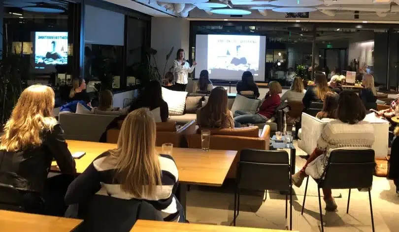 A group of people attend an indoor presentation in a modern office space. They sit on couches and chairs facing a speaker standing near a large screen displaying a slide. The space is well-lit, with large windows and a cozy, professional atmosphere. The focus is on learning and discussion.