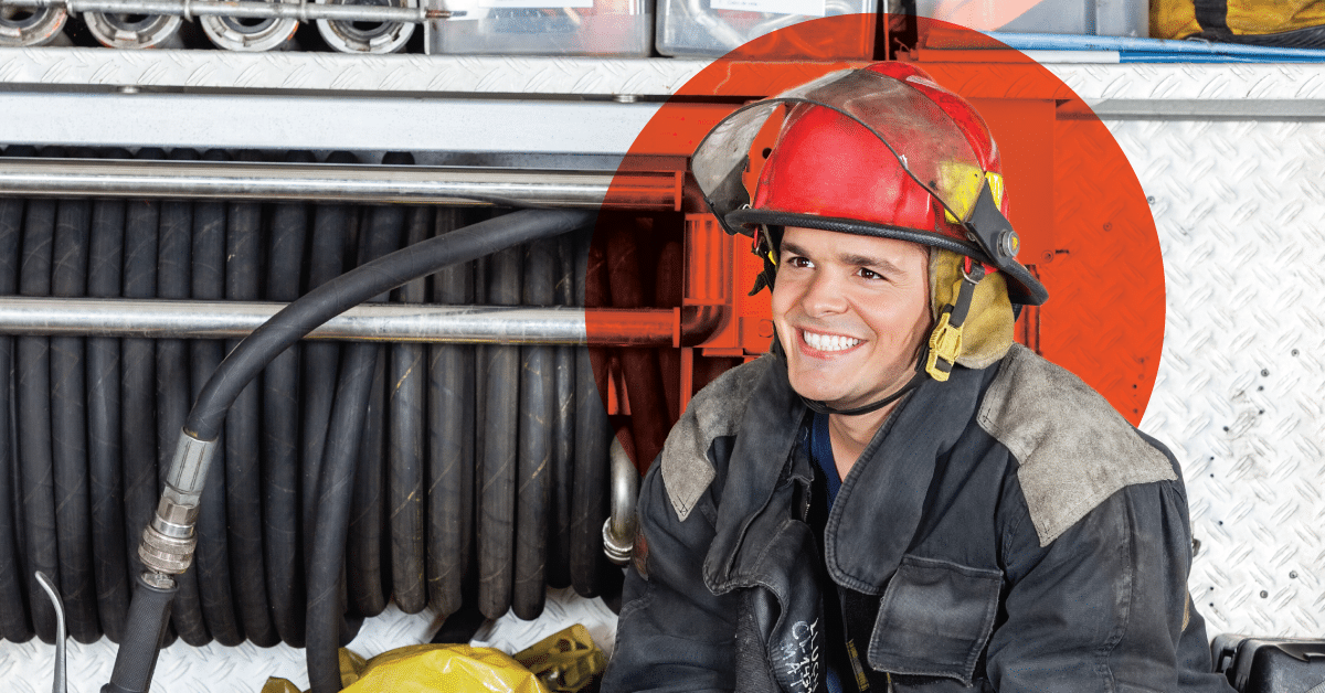 A smiling firefighter wearing a red and yellow helmet and protective gear stands in front of an organized wall of firefighting hoses and equipment. The background features diamond-plated metal, and the firefighter appears to be taking a break from action.