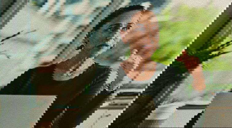 A woman sits at a desk with a laptop, gesturing as she speaks. There is a coffee cup nearby and large windows with a view of trees and buildings in the background.