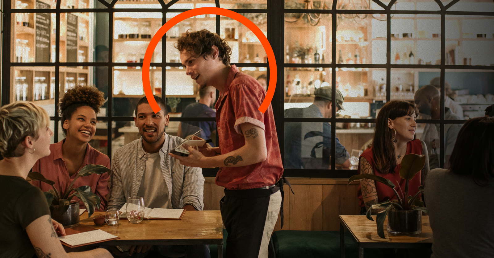 A waiter in a red shirt is taking an order at a restaurant table with three smiling diners; other patrons sit in the background. An orange circle highlights the waiter.