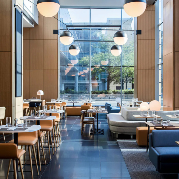 Modern restaurant interior with floor-to-ceiling windows, wooden walls, round ceiling lights, set tables, tan chairs, and lounge seating. City buildings and trees visible outside.