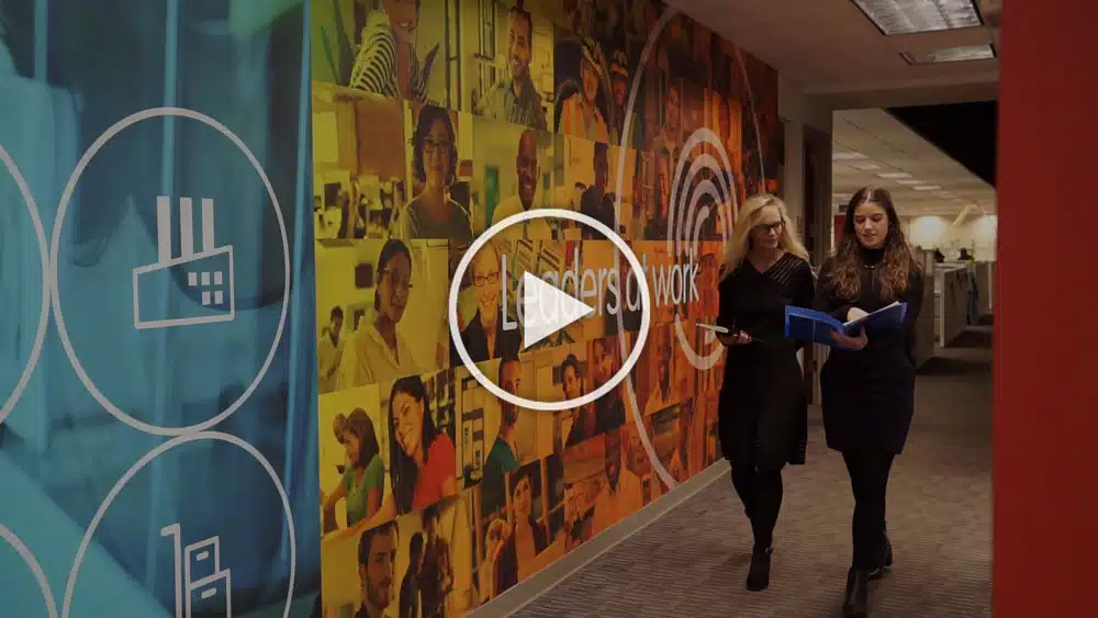 Two individuals walk down a colorful office corridor with graphics and photos on the walls, holding documents.