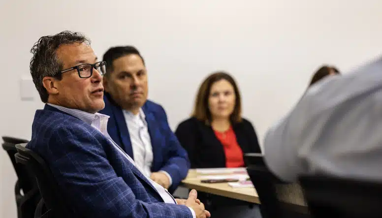 A group of four people are seated at a conference table engaged in discussion. The person in the foreground wears glasses and a blue blazer, while the others listen attentively.