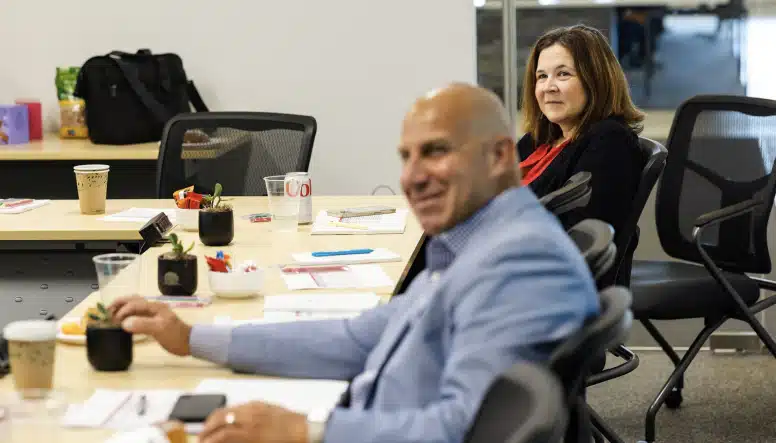 Two people are seated at a conference table, one in focus and smiling while the other, in the foreground, is slightly blurred. The table has various office supplies, drinks, and snacks on it.
