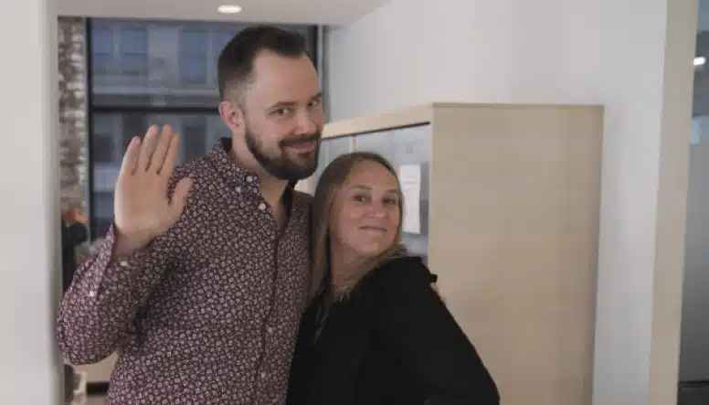 A man and woman stand closely together in an office setting. The man is waving at the camera, while the woman smiles.