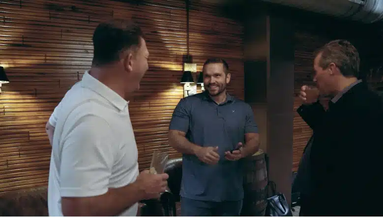 Three men stand in a room with wooden walls, engaged in conversation. One man in a white shirt laughs, a man in a blue shirt smiles, and a man in a black shirt takes a sip from a glass.