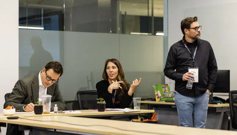 Three people are in a modern office meeting room. One person is writing, another is gesturing, and the third is standing holding a water bottle.