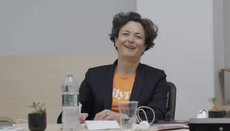 A person wearing a dark blazer and an orange shirt sits at a desk with a smile. The desk has a water bottle, several small plants, a notebook, and various other items.
