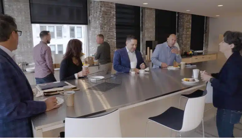 A group of six people gather around a long kitchen counter with drinks and snacks. They appear to be engaging in conversation in an office setting.