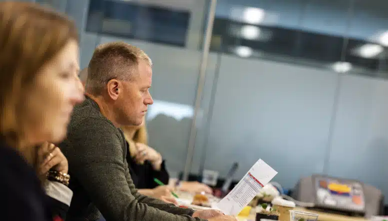 A man in a green sweater reads a document during a meeting, sitting next to a woman in a blurred foreground. The background shows an office setting with glass partitions and lights.