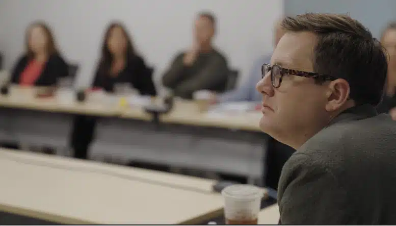 A male participant wearing glasses listens attentively during a meeting, with several other individuals blurred in the background sitting at a conference table.