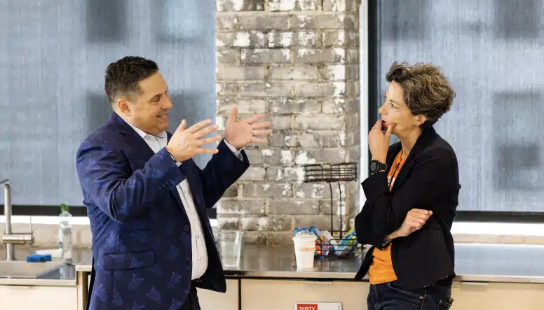 Two people are engaged in a conversation in a modern kitchen area with large windows, exposed brick walls, and a countertop with various items, including chips and a sanitizer bottle.