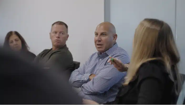 A group of four people seated in a meeting room, engaged in a discussion. One person is speaking while others listen attentively.
