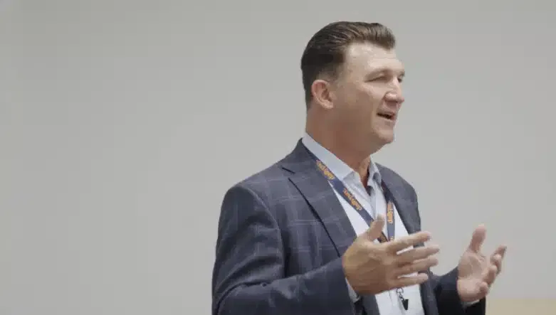 A man in a blue plaid suit speaks while gesturing with his hands. He wears a lanyard around his neck and stands against a plain background.