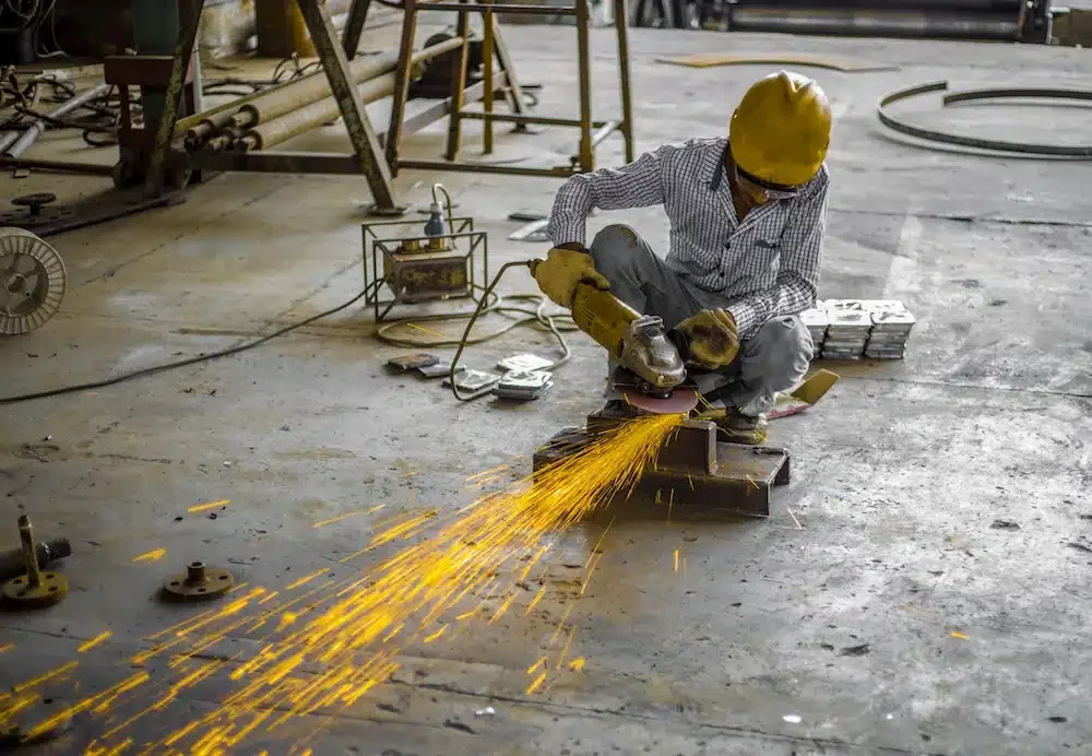 A worker in a yellow hard hat and gloves operates an angle grinder, creating sparks, in a bustling workshop with scattered tools and equipment, highlighting the hands-on skills that can help reduce employee attrition.