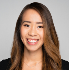 A woman with long, straight hair and a bright smile, wearing a black top and a necklace, against a plain background.