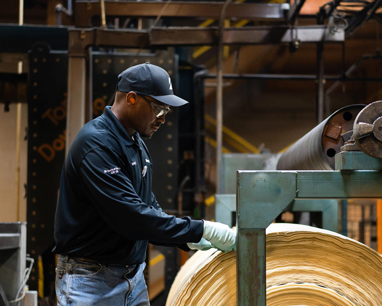 A worker wearing safety glasses, gloves, and a cap inspects a large roll of paper in an industrial facility.