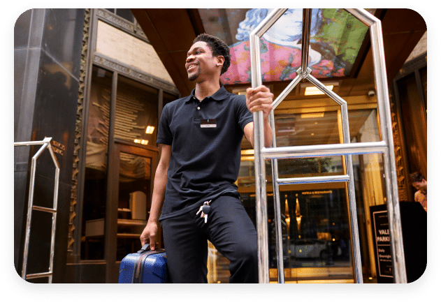 A hotel bellhop, wearing a black shirt and black pants, smiles while leaning on a metal luggage cart. He holds a blue suitcase in his left hand. The background features an elegantly decorated hotel entrance with glass doors and signs, including one for valet parking. With such hospitality charm, it’s clear this place never needs an employee turnover calculator!