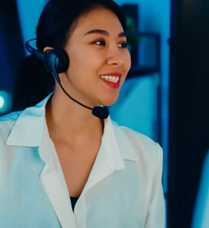 A person with a headset microphone, wearing a white shirt, smiles while looking to the side in a tech-lit environment.