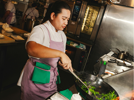 A chef in a white shirt and purple apron stirs vegetables in a wok in a commercial kitchen.