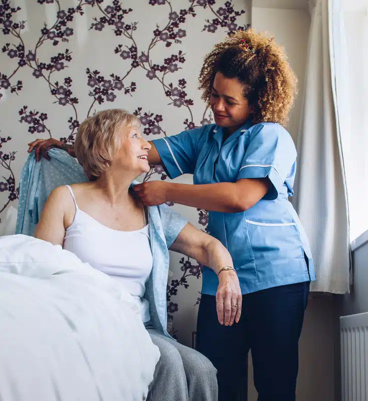 A female caregiver in a blue uniform helps an elderly woman put on a light blue cardigan in a bedroom with floral wallpaper.