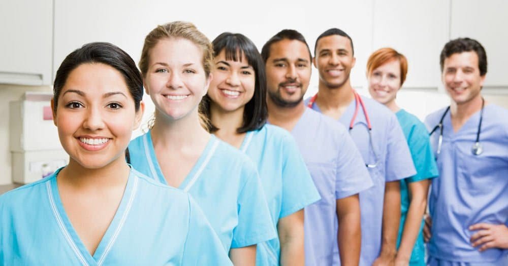 A diverse group of seven smiling healthcare professionals in scrubs and lab coats, standing confidently in a clinical setting.