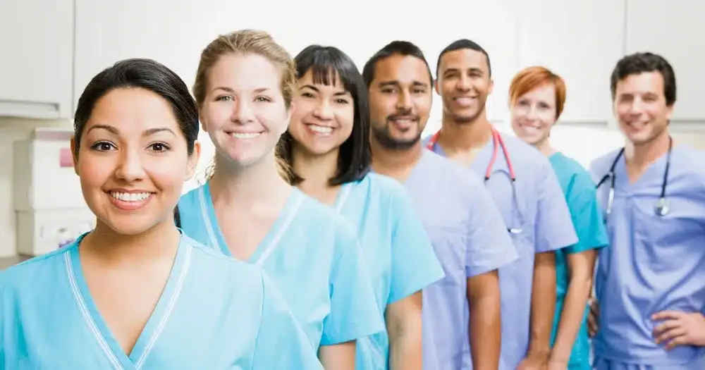 A diverse group of healthcare professionals in scrubs stands in a line, smiling in a medical setting.