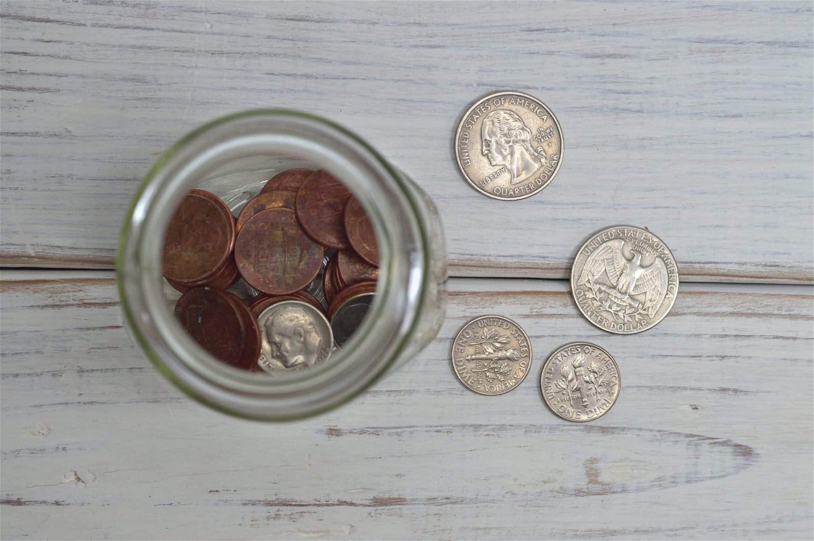 Top view of a glass jar filled with copper pennies next to various silver coins on a wooden surface.