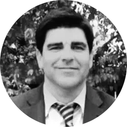 Black and white photo of a man in a suit and tie, smiling, with trees in the background.