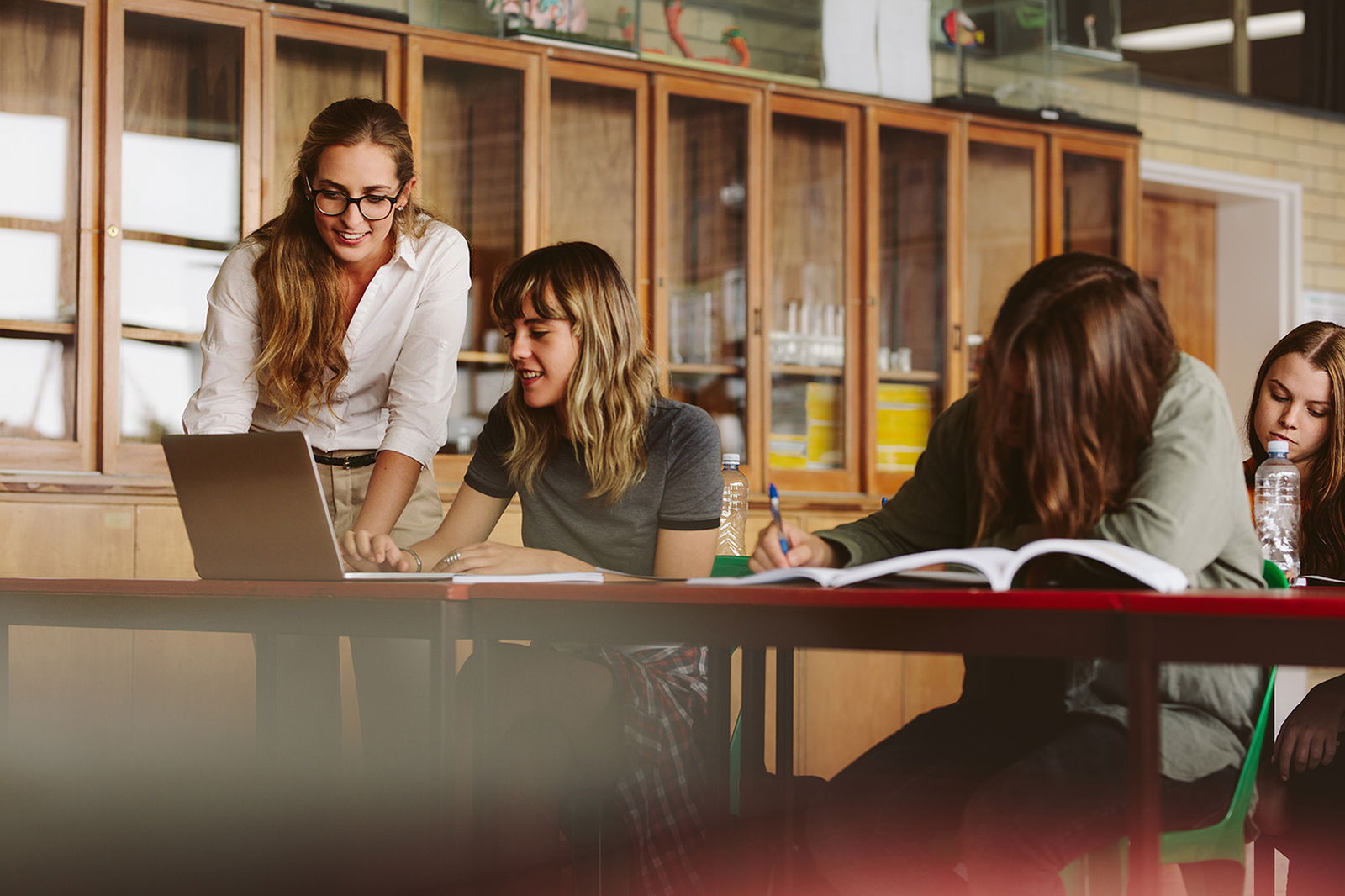 A teacher assists a student working on a laptop while other students write in notebooks at a classroom table.