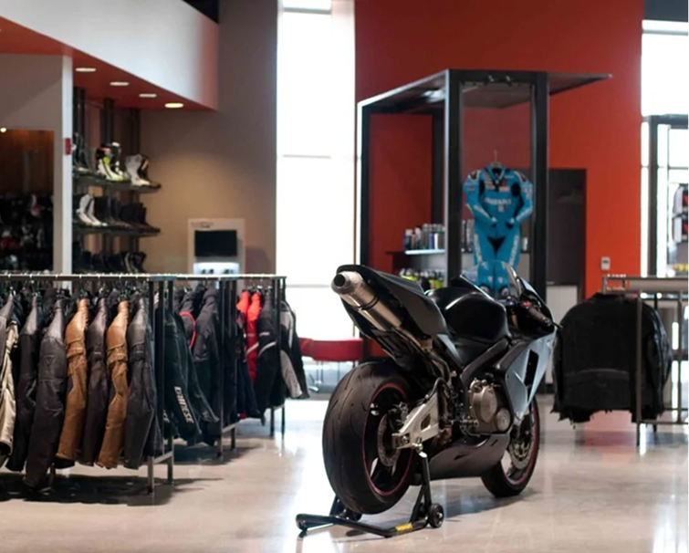 A black sport motorcycle on display inside a shop, surrounded by racks of riding jackets and motorcycle gear.