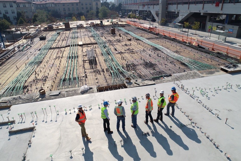 Aerial view of a construction site with a group of workers in safety vests and helmets standing in a line on a large concrete foundation with exposed rebar.
