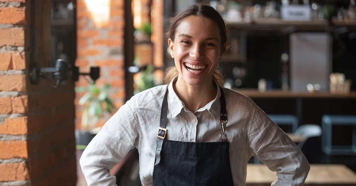 A young woman wearing a black apron and white shirt stands smiling in a rustic-style café with exposed brick walls. She has her hands on her hips and appears cheerful and welcoming. The background includes shelves, plants, and blurred café equipment, indicating a warm and inviting atmosphere.