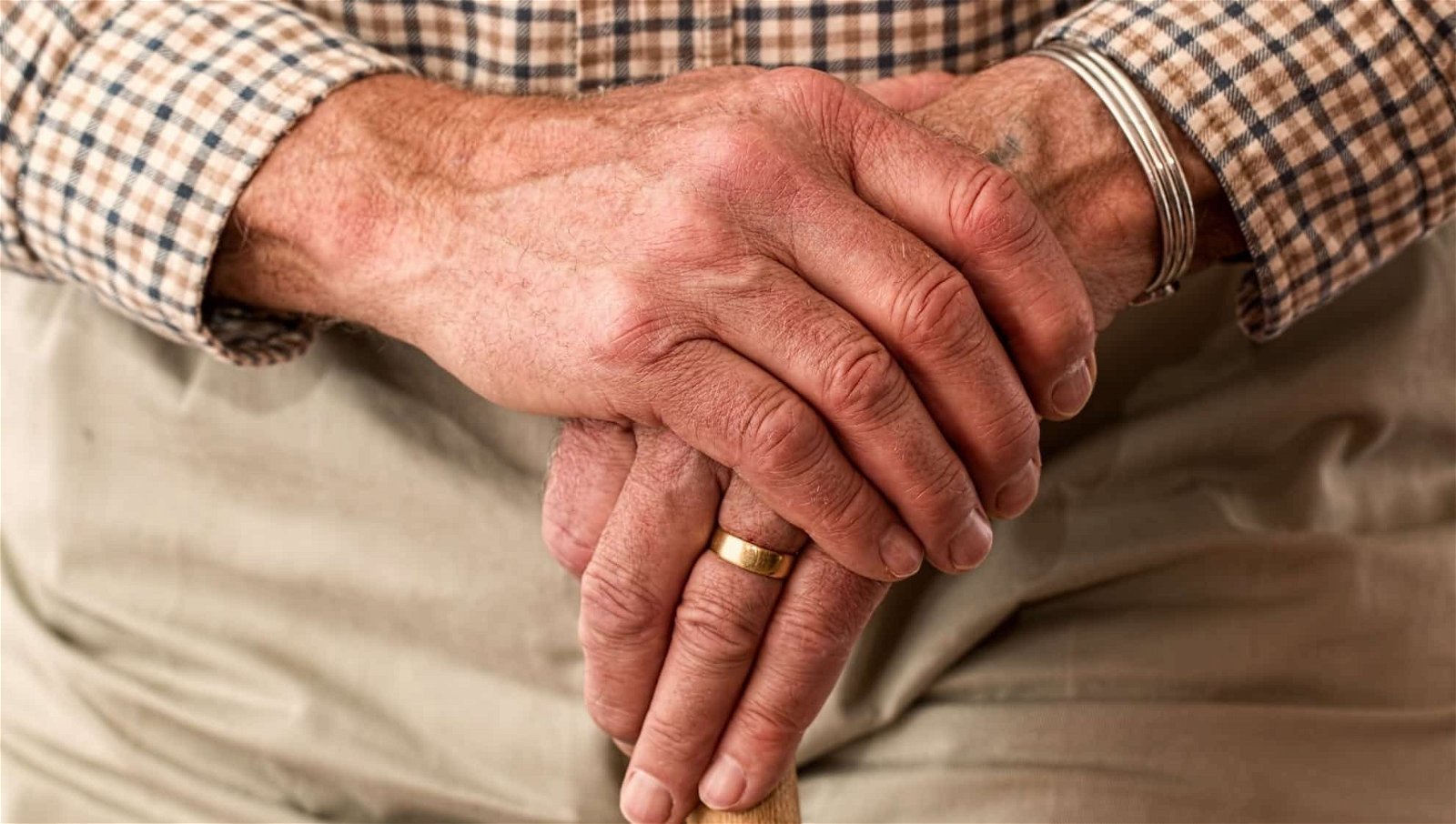 Close-up of an elderly man’s clasped hands resting on his lap, showing details like wrinkles and a wedding ring, highlighting aged skin and timeless love.
