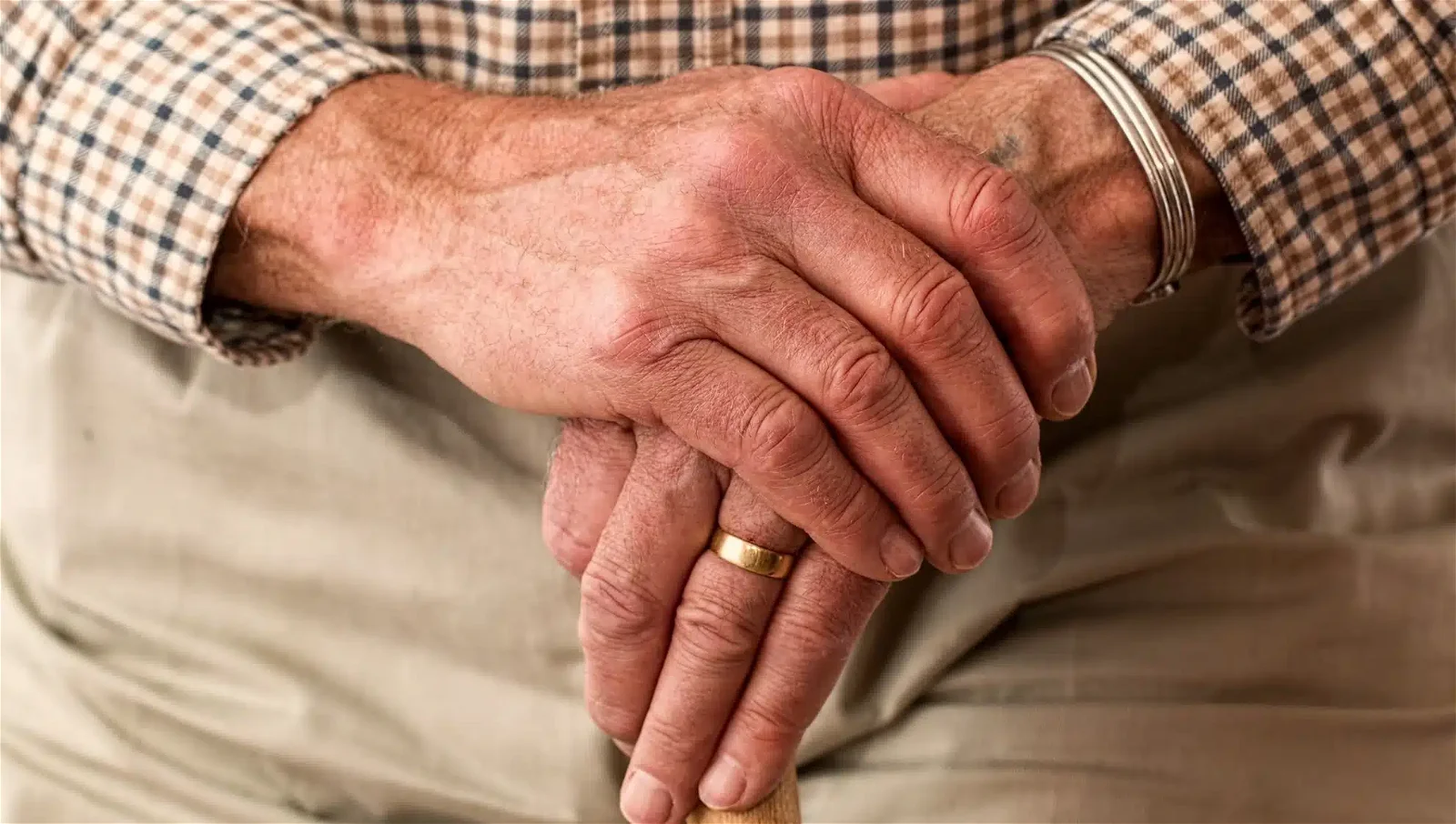 Close-up of elderly hands resting on a cane. The person is wearing a plaid shirt, a ring on one finger, and a bracelet.