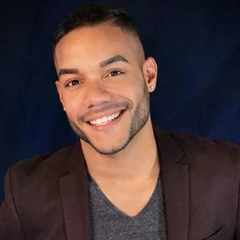 Smiling man with short hair, beard, and earrings wearing a dark blazer and gray shirt against a dark blue background.